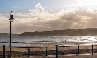 A promenade beside a beach with a headland in the distance and a cloudy sky.