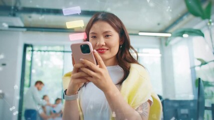 Smiling Asian female holding smartphone and reading messages in bright office. Digital icons floating around while enjoying conversation. Standing near large windows in busy workplace. - Powered by Adobe