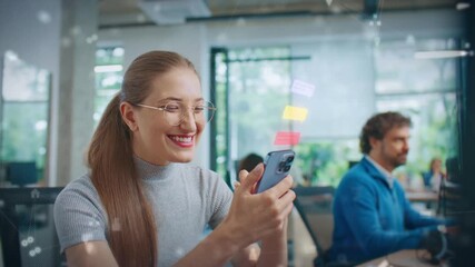 Smiling Caucasian female holding smartphone and reading colorful chat bubbles in bright workplace. Sitting near coworker while enjoying friendly message exchange in modern office environment. - Powered by Adobe