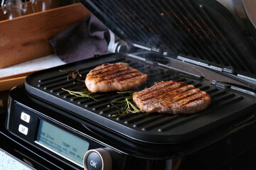 A close-up of marbled beef steaks grilling on an electric grill against the backdrop of a home kitchen. Cooking delicious and healthy food at home.