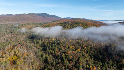 Aerial view of fog-covered Adirondack forest with autumn foliage and distant mountain peaks under clear blue sky.