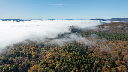 Aerial view of fog-covered Adirondack forest with autumn foliage and distant mountain peaks under clear blue sky.