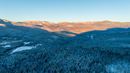 Snow-covered forest and mountain range at sunrise with golden light casting across the winter landscape.