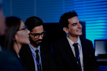 A diverse team with a Caucasian man, Indian man, and Asian woman in a modern tech office. They are collaborating and discussing a project during a late meeting.