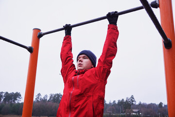 Fototapeta premium Front view of a young athlete hanging from a horizontal bar at a street sports field
