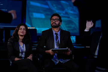 A diverse team with an Indian man and an Indian woman sits in a business briefing. The man holds a tablet, and the woman smiles while they listen to a presentation.