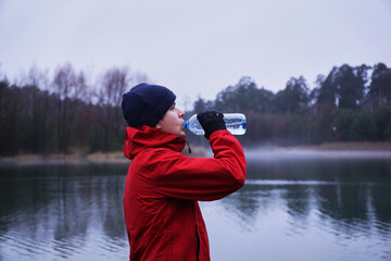 A male athlete drinks water from a bottle during an outdoor workout by a lake. The man is resting after a jog in the park, replenishing his body with water