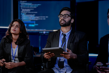 A large, diverse team of business professionals in a group meeting. An Indian man with a tablet leads the discussion with Asian and Caucasian colleagues in a high-tech office.