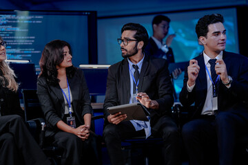 A large, diverse team of business professionals in a group meeting. An Indian man with a tablet leads the discussion with Asian and Caucasian colleagues in a high-tech office.