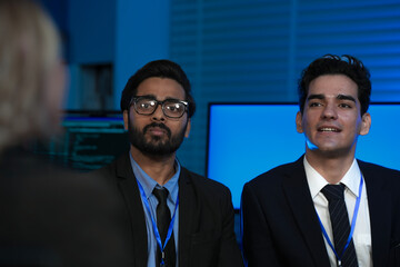 A diverse team, an Indian man with glasses and a Caucasian man, listens intently during a professional business meeting or conference in a modern, high-tech office at night.