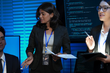 An Indian female developer leads a code review, holding a report. She explains a Python script for an AI or cybersecurity system to her team in a dark SOC Security Operations Center.