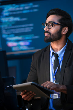 An Indian male software engineer or data scientist holds a tablet in a command center. He is asking a question about the AI code or cybersecurity data shown on the large screen.