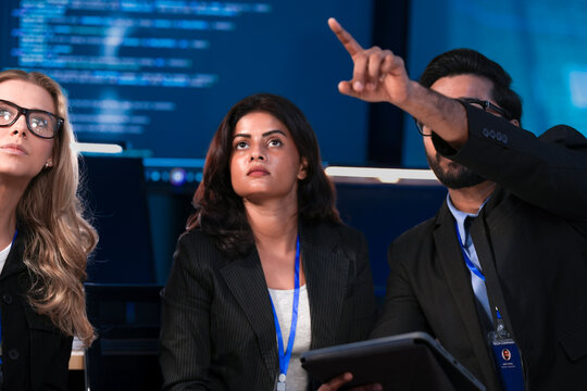 An Indian data scientist explains an AI algorithm on a tablet to a Caucasian software engineer. They are reviewing system data in a cybersecurity command center