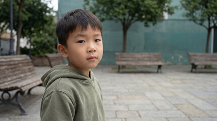 Thoughtful young boy in green hoodie standing alone in quiet urban park with empty benches and trees in soft daylight