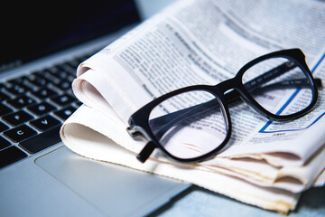 A pile of newspapers ,glasses and computer on the table.