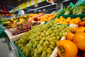 Fresh fruits are displayed in baskets at a grocery store. The scene features green grapes, oranges.