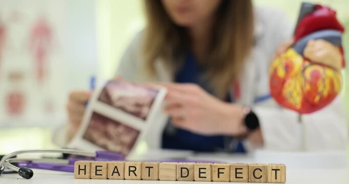 Experienced woman cardiologist carefully examines printed ultrasound images near heart model. Wooden cubes on table clearly form phrase Heart Defect