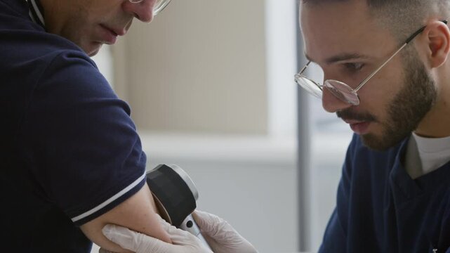 Focused young Middle-Eastern male dermatologist in scrubs and sterile gloves examining middle-aged Caucasian male patients arm, looking at his skin with dermatoscope and asking questions