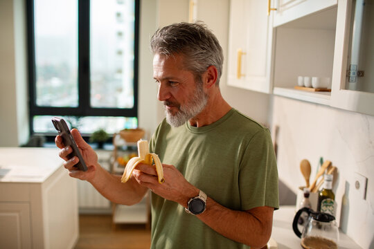 Mature man focused on smartphone while eating banana in home kitchen