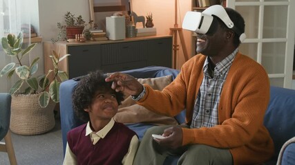 Senior African American grandfather immersed in VR gaming with teenage grandson helping granddad exploring metaverse sitting on couch in cozy living room - Powered by Adobe