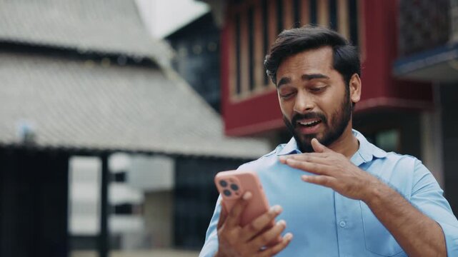 Portrait of friendly Indian businessman talks with sign language on video call outdoors slow motion. Man gestures and gives thumbs up via chat on smartphone at urban block