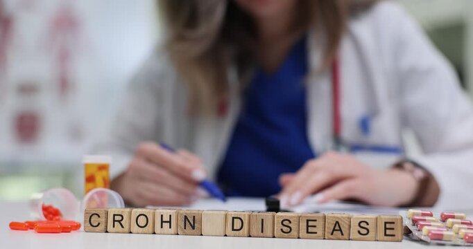 Woman physician sits at desk and analyzes medical history of patient. Wooden cubes neatly arranged in row form phrase Crohn Disease near pills