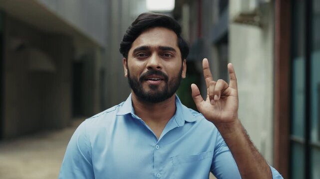 Portrait of smiling Indian interpreter in shirt uses sign language standing in city district. Businessman saying Hello, I love you so much in an urban street environment.