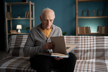 Lonely elderly widower sitting on couch looking at old photos recalling memories of deceased wife