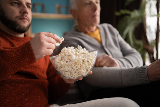 Close-up of senior father and middle-aged son eating popcorn watching television together at home