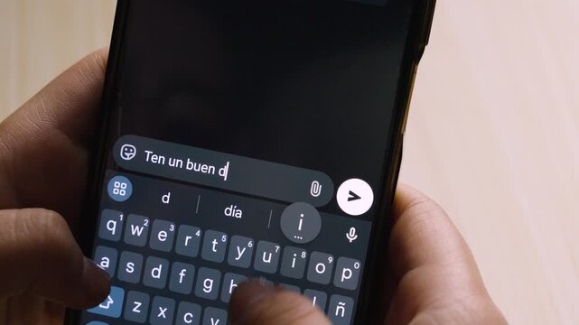 Close-up of a person holding a smartphone while typing and preparing to send the message &ldquo;Ten un buen d&iacute;a :)&rdquo; in a chat application. The phone&rsquo;s keyboard and send button are clearly visible.