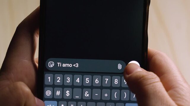 Close-up of hands typing &ldquo;Ti amo <3&rdquo; on a smartphone messaging app, showing a dark chat interface and touchscreen keyboard in use.