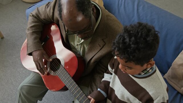 High angle shot of little Black boy pressing strings on guitar fretboard exploring musical instrument with grandfather in living room, family connecting through music