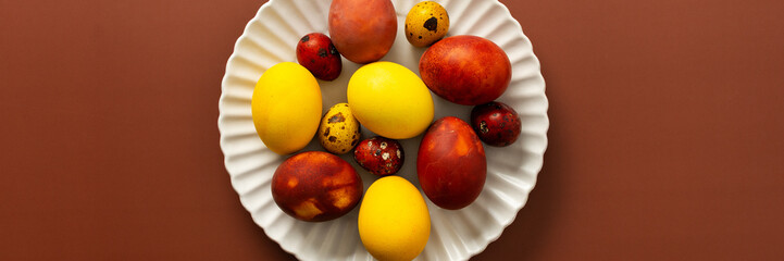 A collection of brightly colored Easter eggs in various sizes and patterns is displayed on a white scalloped plate, resting on a warm brown surface, banner