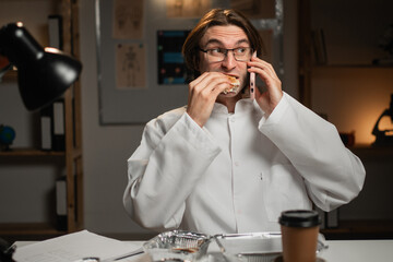 Exhausted male doctor calling by phone during break for takeout meal at night shift