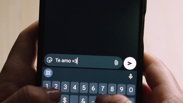 A hand holding a smartphone, typing a romantic message ("Te amo <3") in Spanish on a dark screen, symbolizing digital love, chat, and affection.