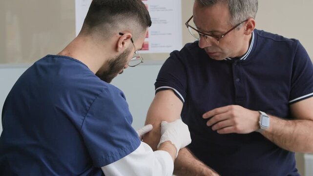 Tilt down shot of middle-aged Caucasian male patient sitting in doctors office and showing skin on forearm to young male dermatologist in scrubs and sterile gloves examining carefully