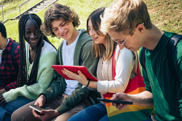 Diverse group of teenage students sitting outdoors sharing digital tablets and smartphones on sunny day