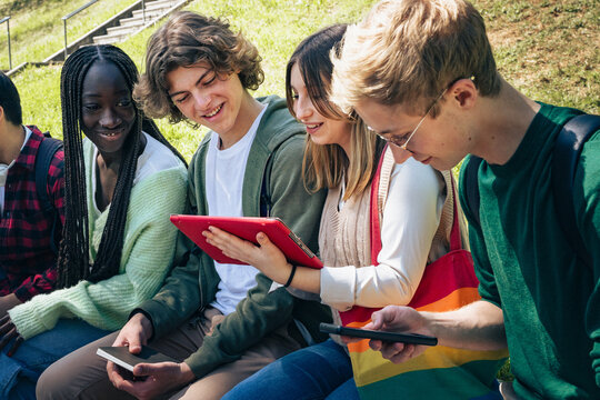 Diverse group of teenage students sitting outdoors sharing digital tablets and smartphones on sunny day - Powered by Adobe