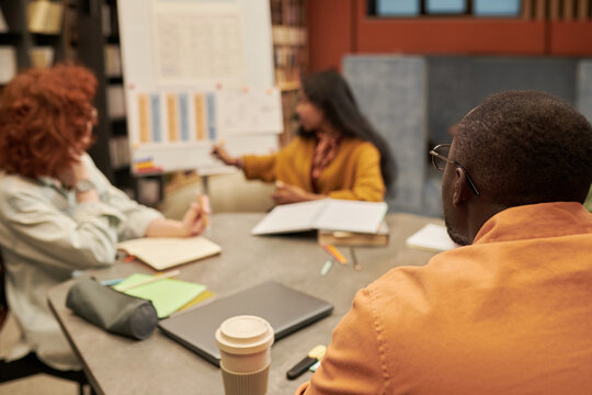 Diverse group of young people collaborating around table, Black man and woman listening while Asian woman presenting data on flip chart during meeting in office