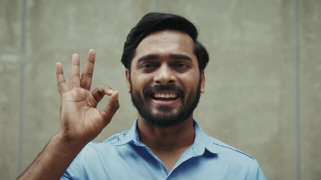 Portrait of smiling Indian man shows Ok gesture as stands near grey wall. Happy Hindu businessman expresses accept with common hand sign outdoors. Body language communication