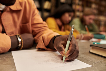 Black young man writing on paper at desk in library with multiethnic young people studying in background, focusing on hand holding pen and academic environment