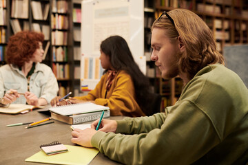 Young man writing notes while sitting at table with multiethnic young women studying together in library, textbooks and notebooks on desk