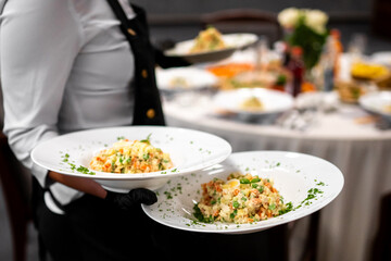 A waiter in a formal uniform is serving plates of a creamy vegetable salad, possibly a Russian or Olivier salad, in a restaurant.
