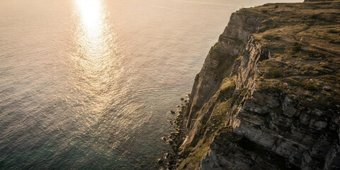 Aerial view of a massive rocky cliff towering over a calm ocean or sea in warm golden light.