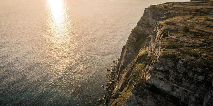 Aerial view of a massive rocky cliff towering over a calm ocean or sea in warm golden light. - Powered by Adobe