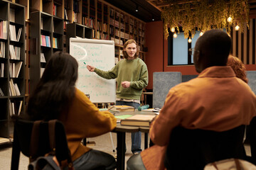 Young man standing at whiteboard teaching language lesson to diverse group of young people seated at table in library setting, students listening and taking notes