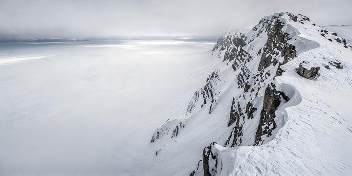 A rugged, snow-covered winter mountain ridge providing a panoramic view over a snowy valley or frozen lake.