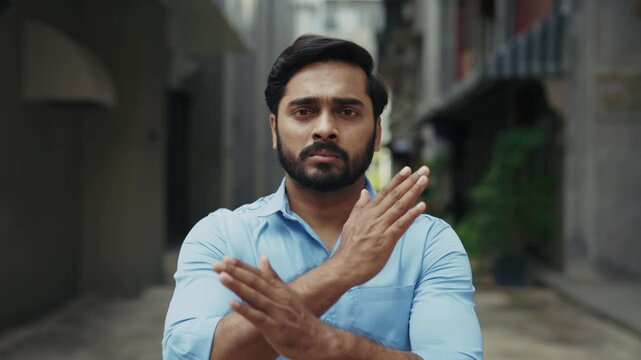 Portrait of serious Indian man shakes head and gives Stop sign by hands on street. Worried Hindu male in blue shirt shows No gesture for active protest in old town district