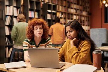 Young woman and young Black woman sitting at table using laptop together in library, studying or collaborating on project, books and papers scattered on desk