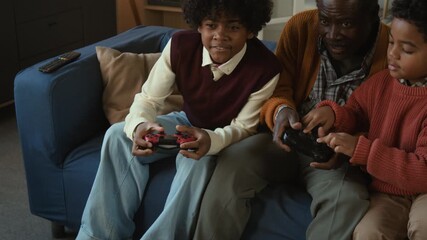 Cheerful teenage Black boy rejoicing at victory in video game on console playing with sibling and grandfather while sitting on couch in living room - Powered by Adobe